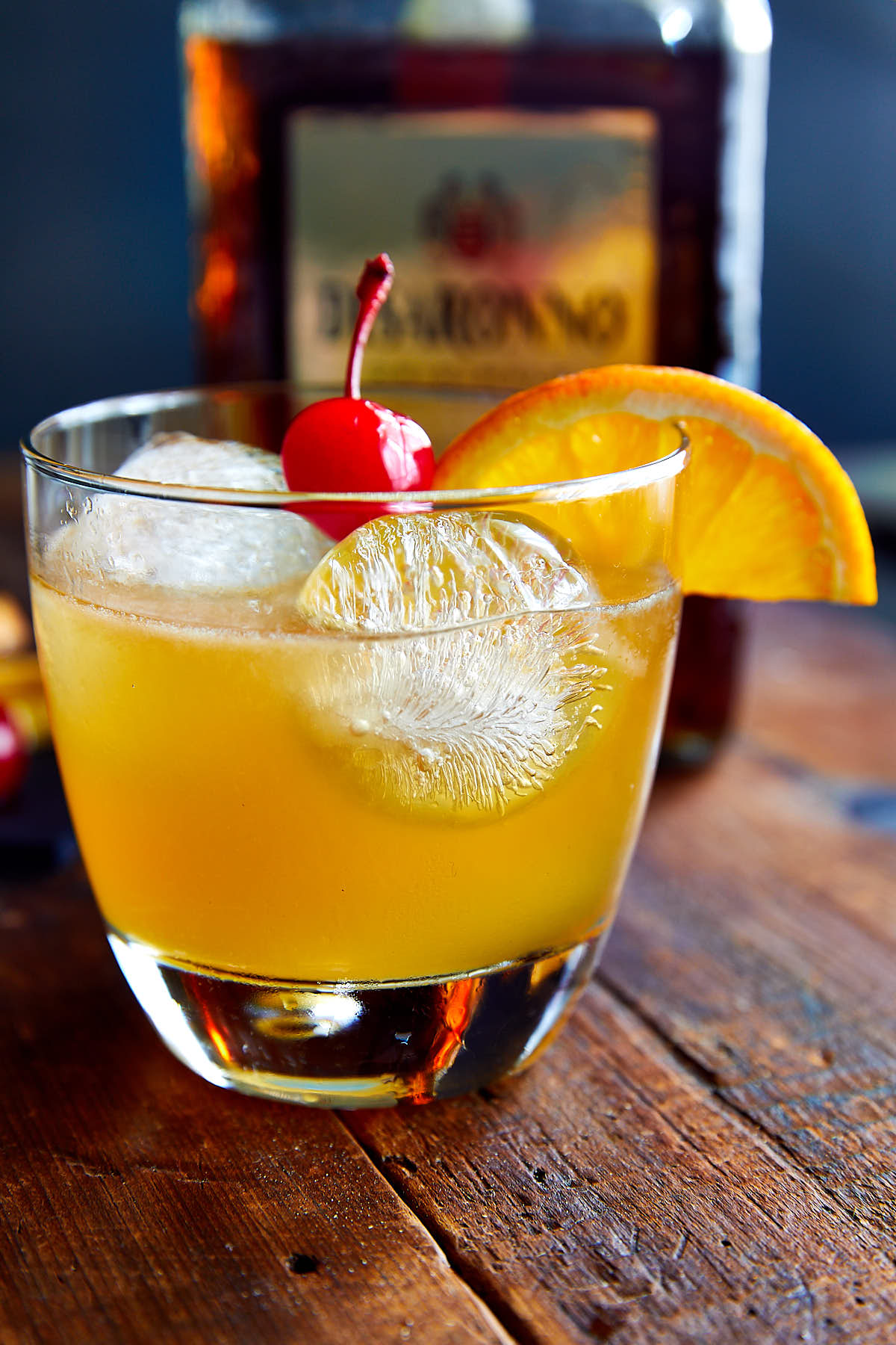 Amaretto sour drink in a glass with ice, garnished with a slice of orange and a maraschino cherry.