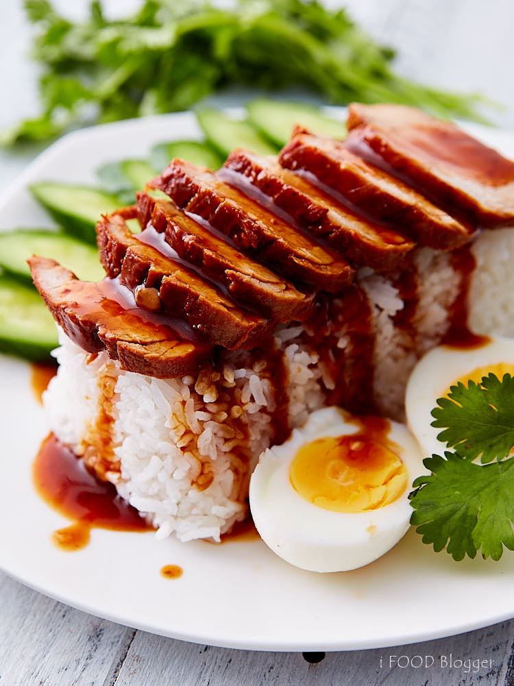 Thai BBQ pork on rice with boiled eggs and cucumbers on a white plate, with sauce poured over.