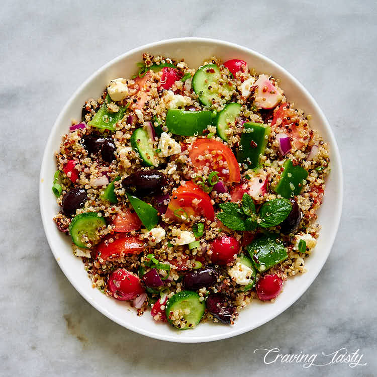 Zesty quinoa salad with chunks of tomatoes, bell pepper, cucumbers and black olives on a white plate.