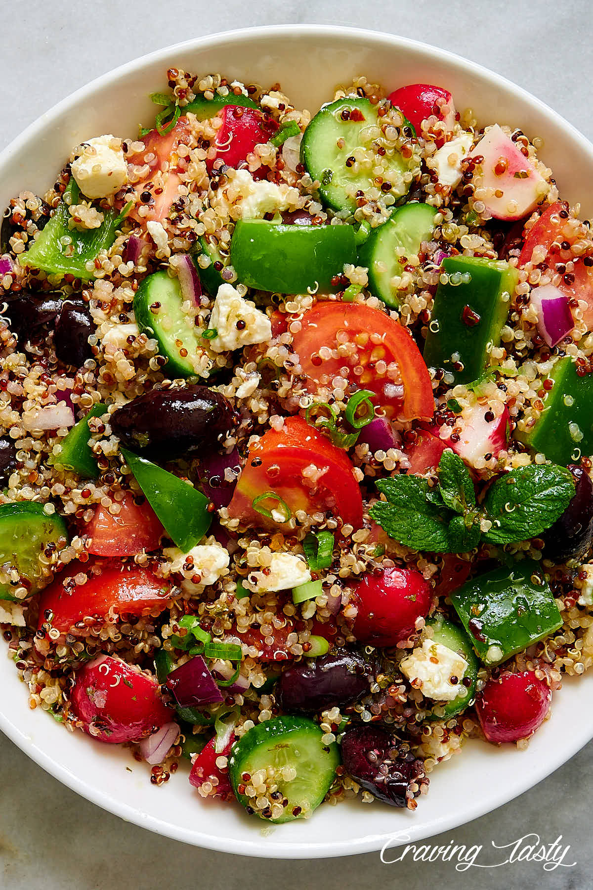 Zesty quinoa salad with chunks of tomatoes, bell pepper, cucumbers and black olives on a white plate.