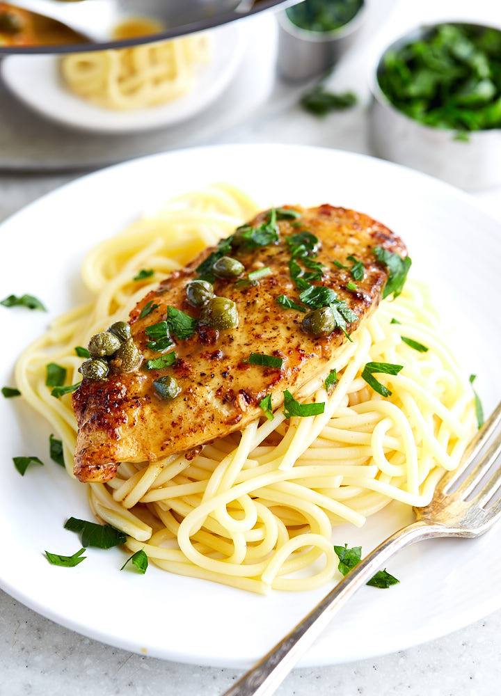 Chicken piccata over a bed of pasta on a white plate, topped with capers and chopped parsley.