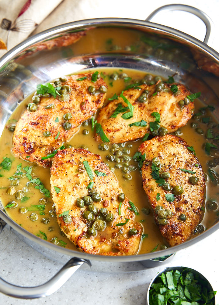 Chicken piccata in a stainless steel saute pan topped with capers and chopped parsley, with juices on the bottom.