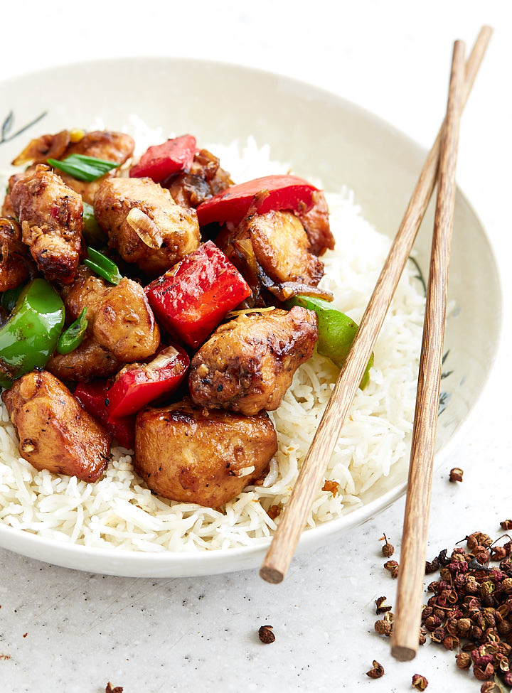 Szechuan chicken in a bowl over white rice with chopsticks on the side.