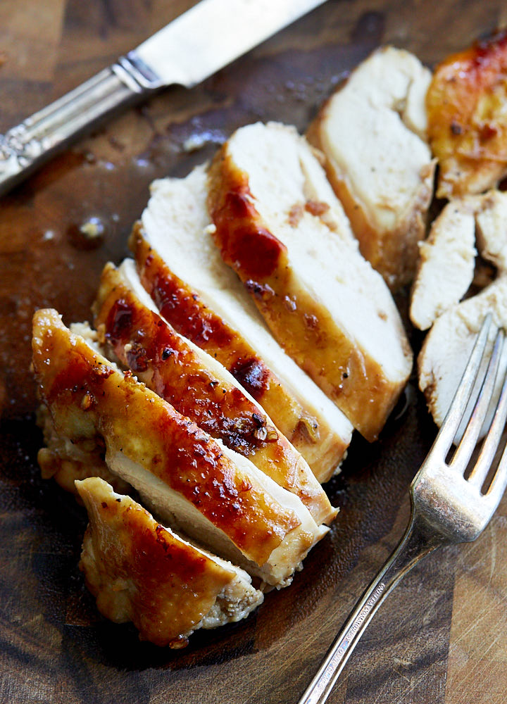 Sliced Asian chicken breast on a brown cutting board with a fork an knife on the side.