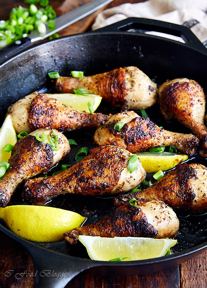 Crispy, well-browned chicken drumsticks on a cast iron pan garnished with lemon wedges and chopped green onions.