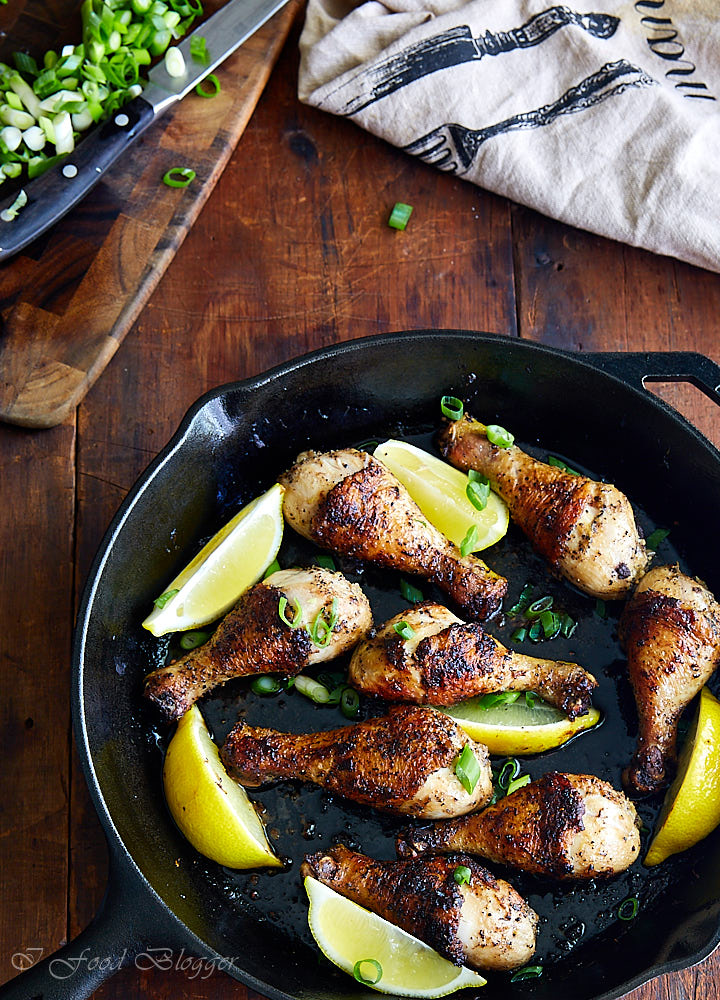 Top down view of pan seared chicken drumsticks in a cast iron pan, cutting board with onions and a towel nearby.