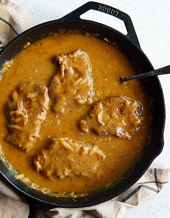 Top down view of a large cast iron pan with pork chops smothered with dark brown sauce and onions.