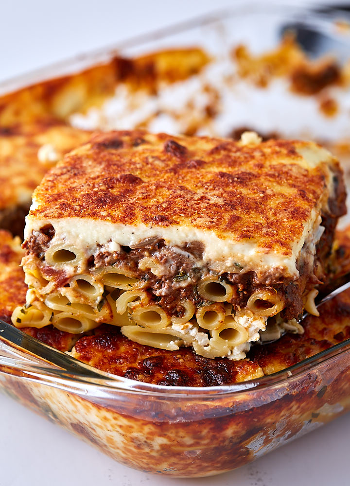 A slice of Greek lasagna on top of lasagna in a baking dish.