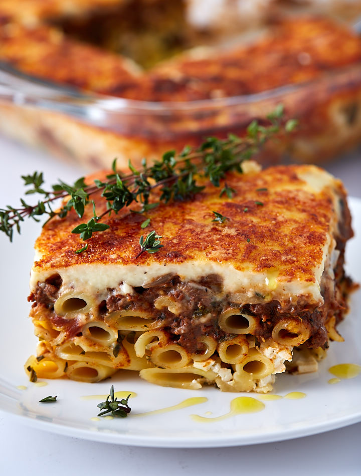 A slice of Greek Lasagna on a white plate drizzled with olive oil and garnished with a twig of Thyme.