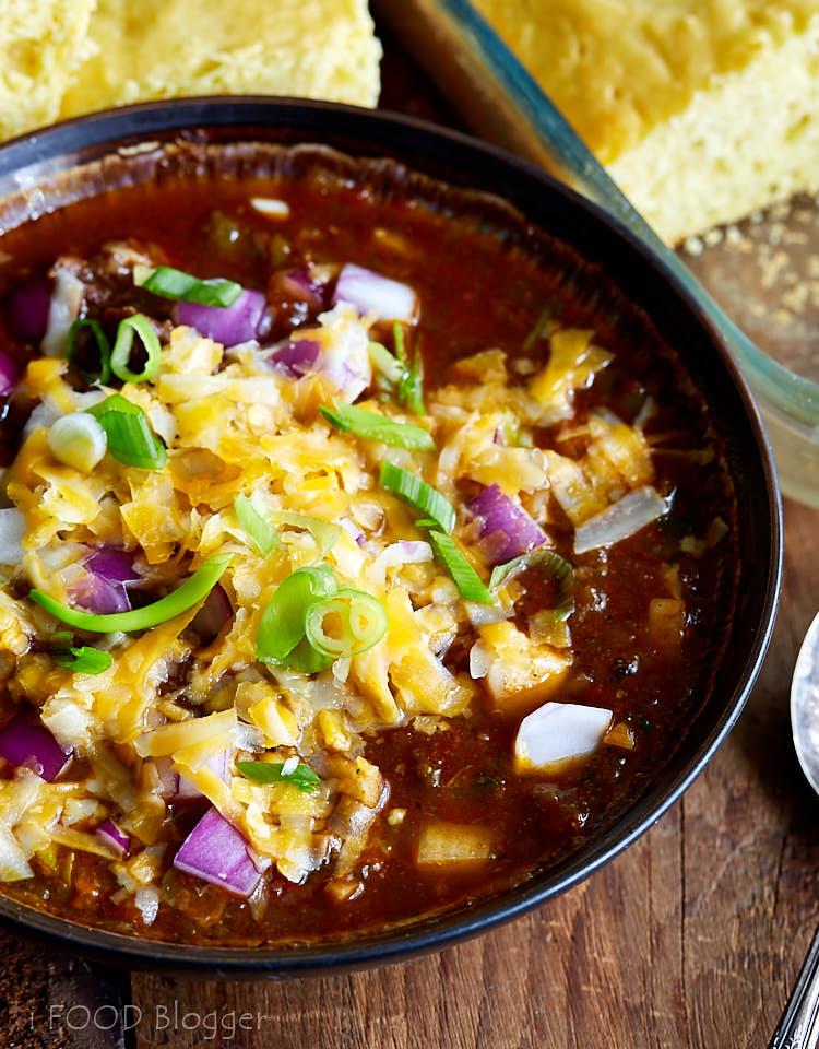 Texas chili garnished with shredded cheddar cheese and chopped onions in a bowl with corn bread next to it.