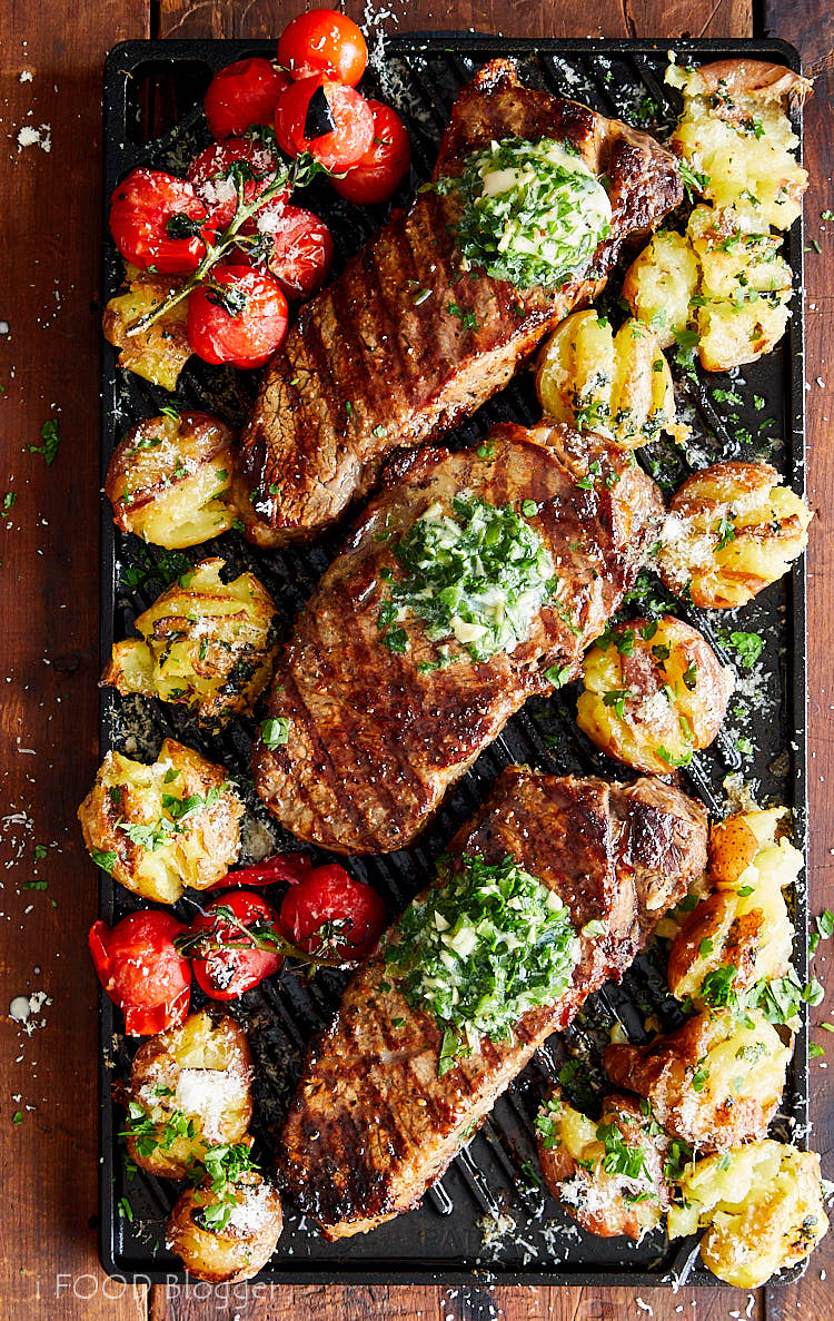 Broiled steaks on a cast iron griddle with smashed potatoes and tomatoes.