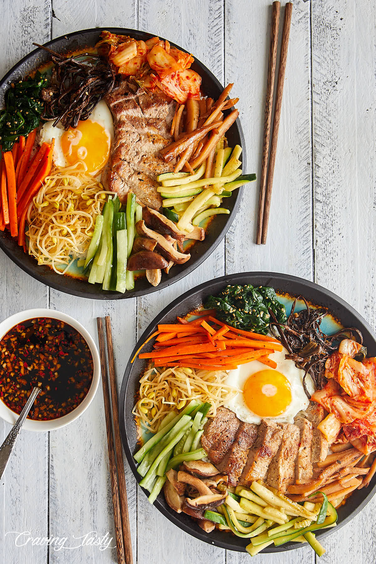 Two bibimbap bowls with a sauce and chopsticks next two them on a white table.