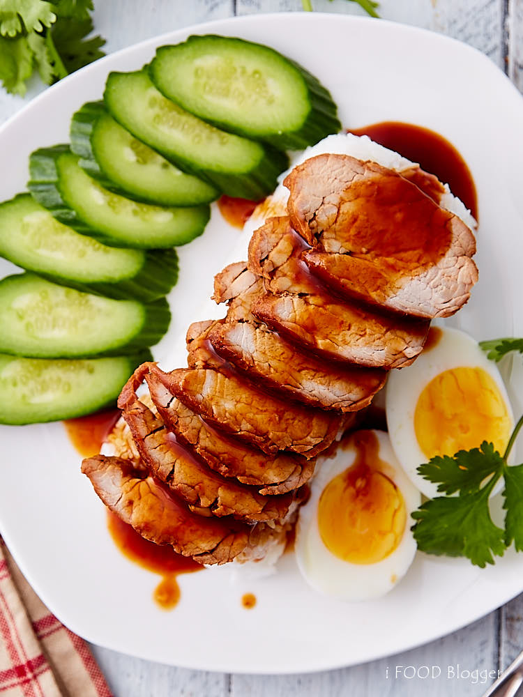 Top down view of Thai BBQ pork on rice with boiled eggs and cucumbers on a white plate, with sauce poured over.