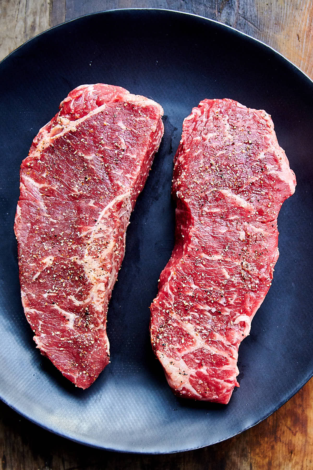 Two beautifully marble raw strip-loin steaks on a black plate.