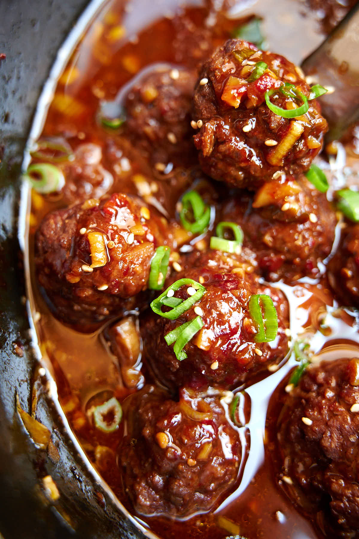 Top down view of Asian meatballs, inside a Crock Pot, soaked in sauce and sprinkled with sliced green onions and sesame seeds.