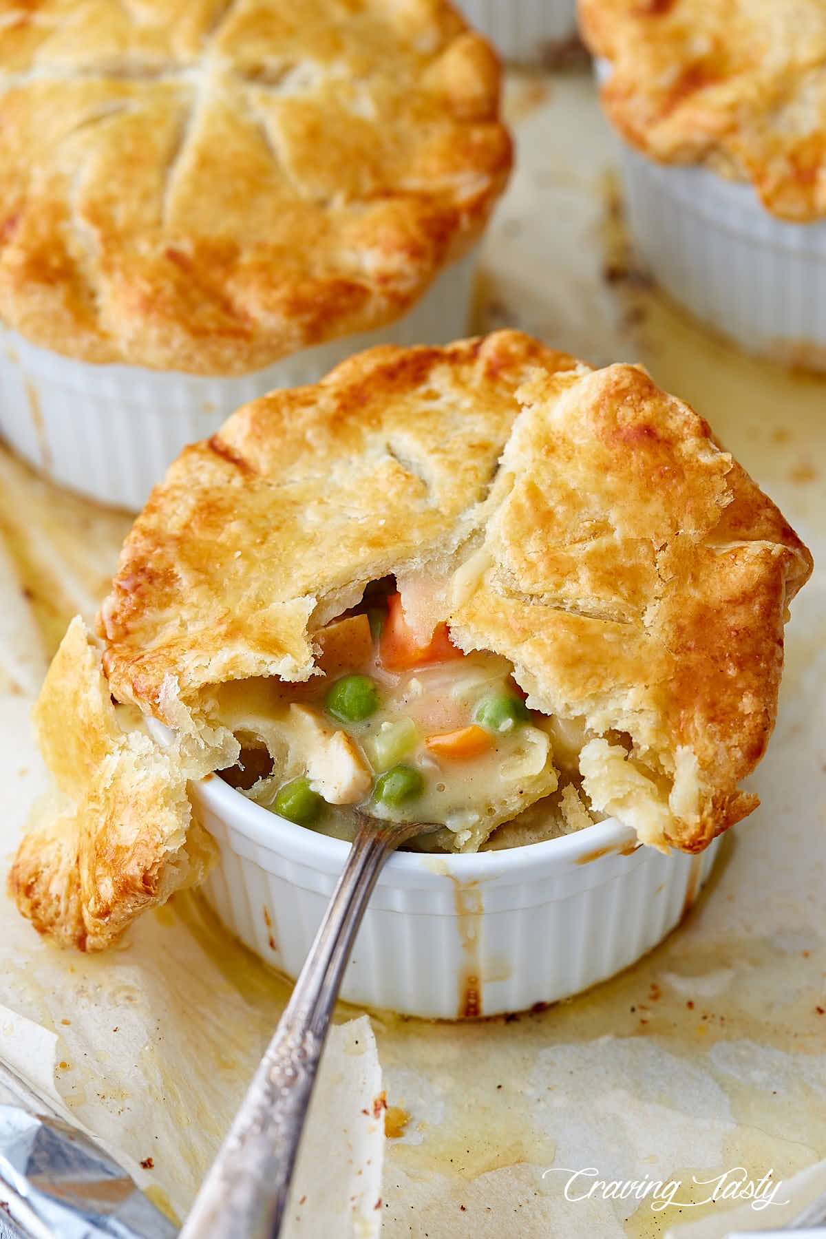 Chicken pot pie, in a ramekin, part of crust removed, a spoon inserted into creamy pie filling, chunks of chicken, peas and carrots visible.