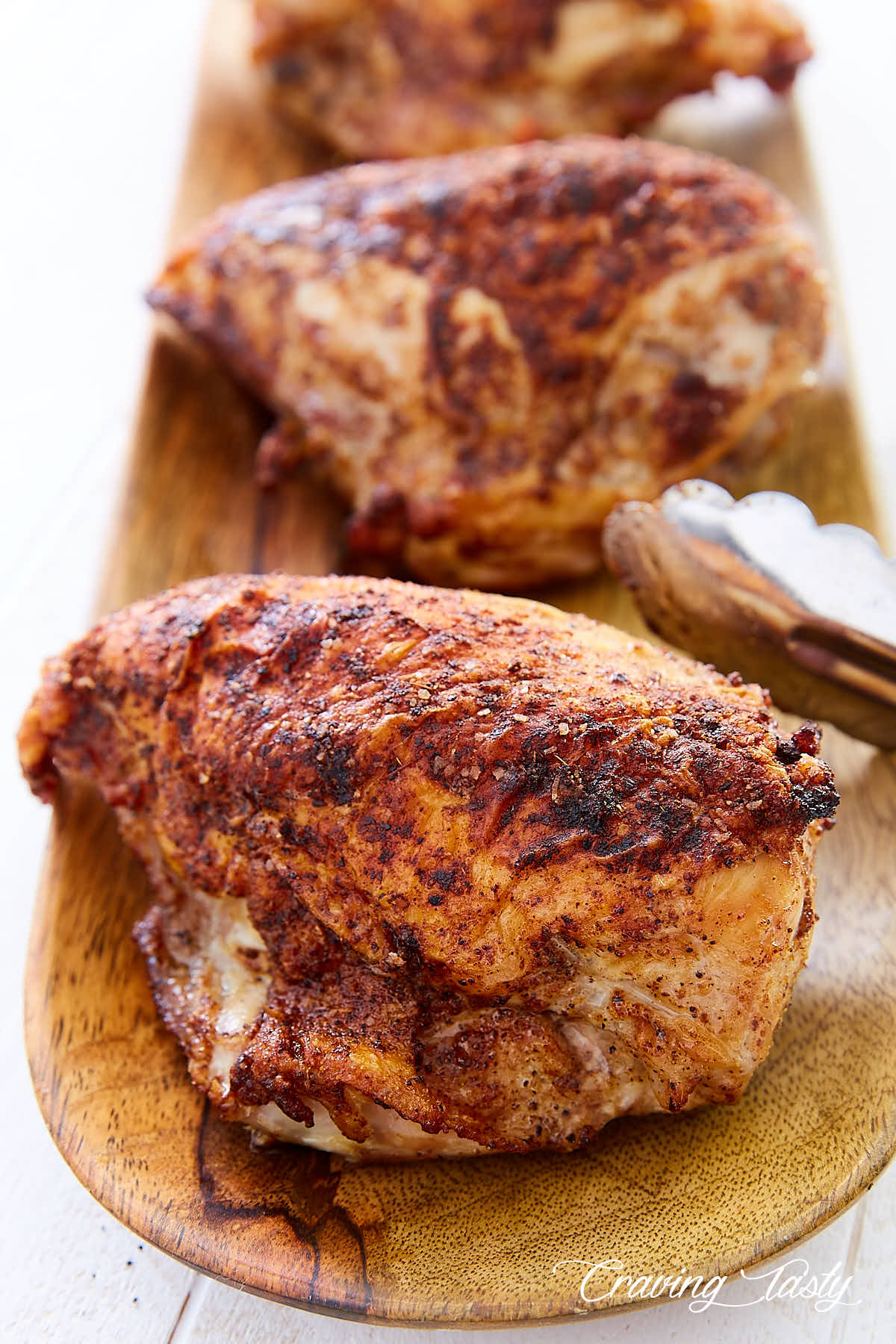 A close up of a crispy baked chicken breast on a wooden plate with more chicken breasts on the background.