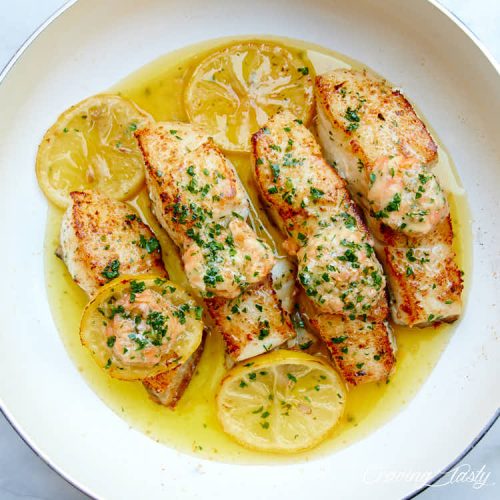 Seared halibut filets in oil in a white pan, with lemon slices .