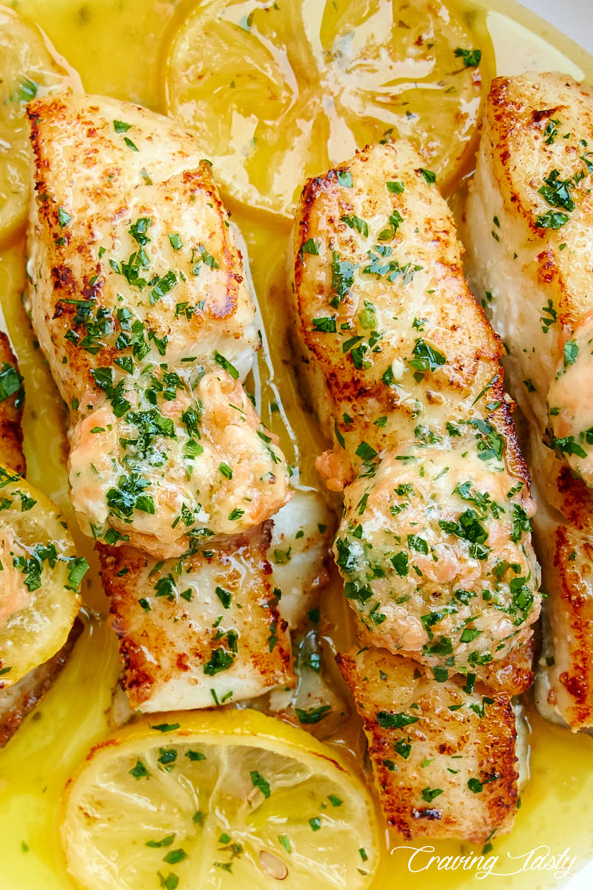 Close up of browned halibut pieces in oil, topped with salmon butter and chopped parsley.