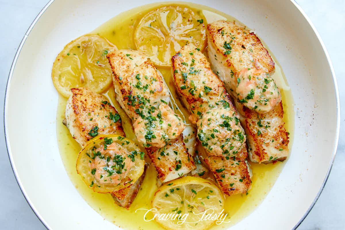 Four well browned halibut steaks in a white pan with oil, lemon slices and salmon butter on top.