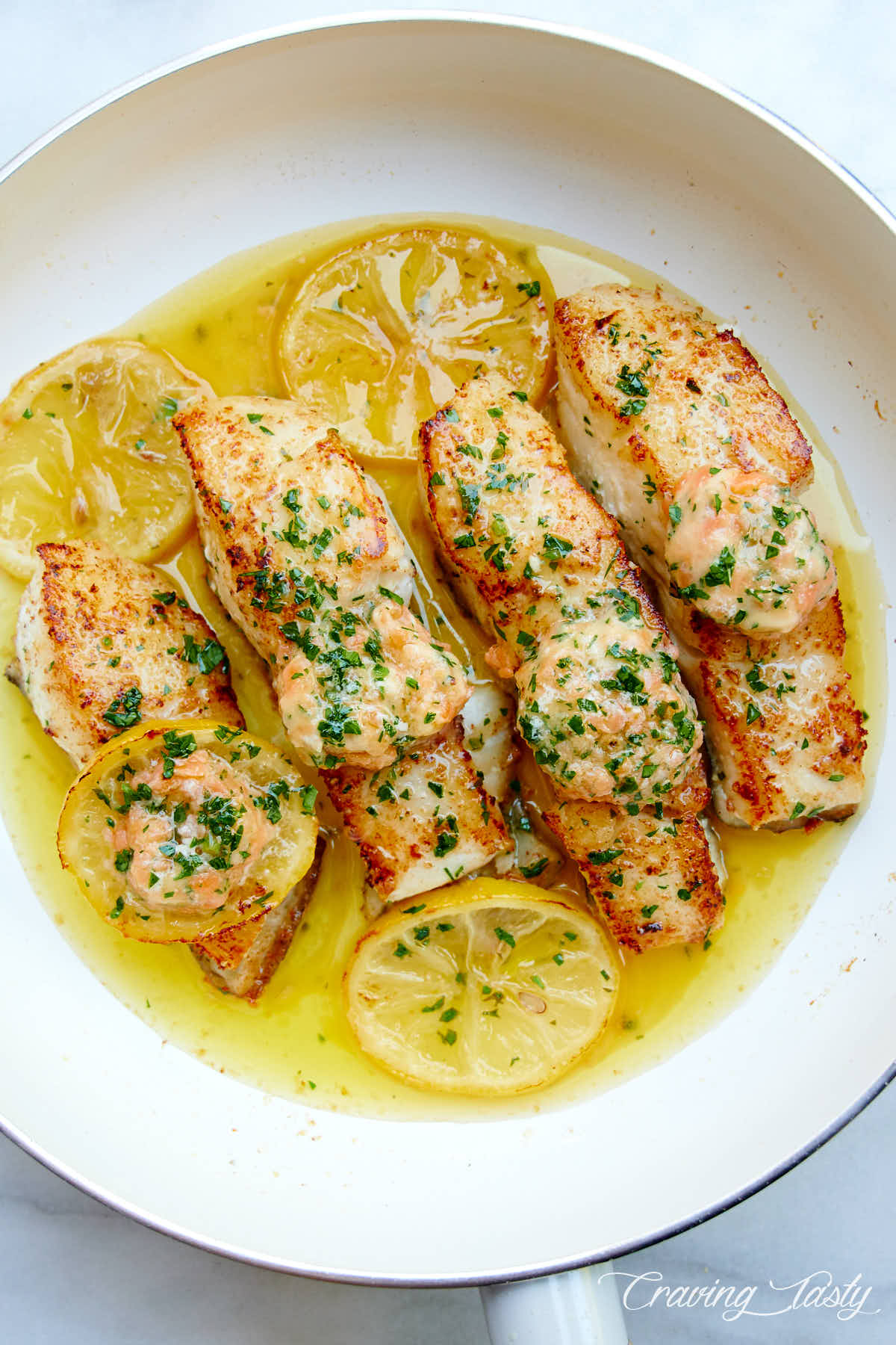 Seared halibut pieces in oil in a white pan, with lemon slices .