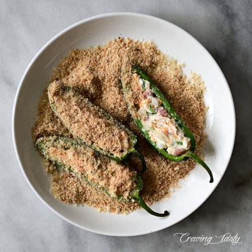 Stuffed raw jalapeno peppers dredged in bread crumb and Parmesan cheese mix in a shallow bowl.