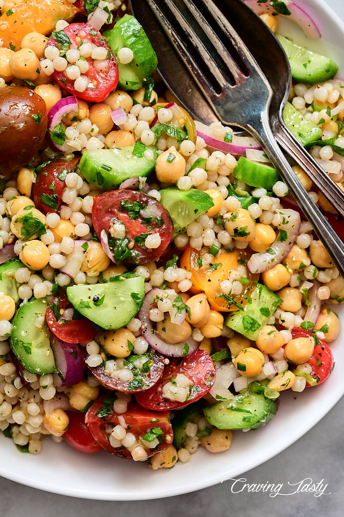 Israeli couscous salad in a white bowl with a fork and a spoon inside.