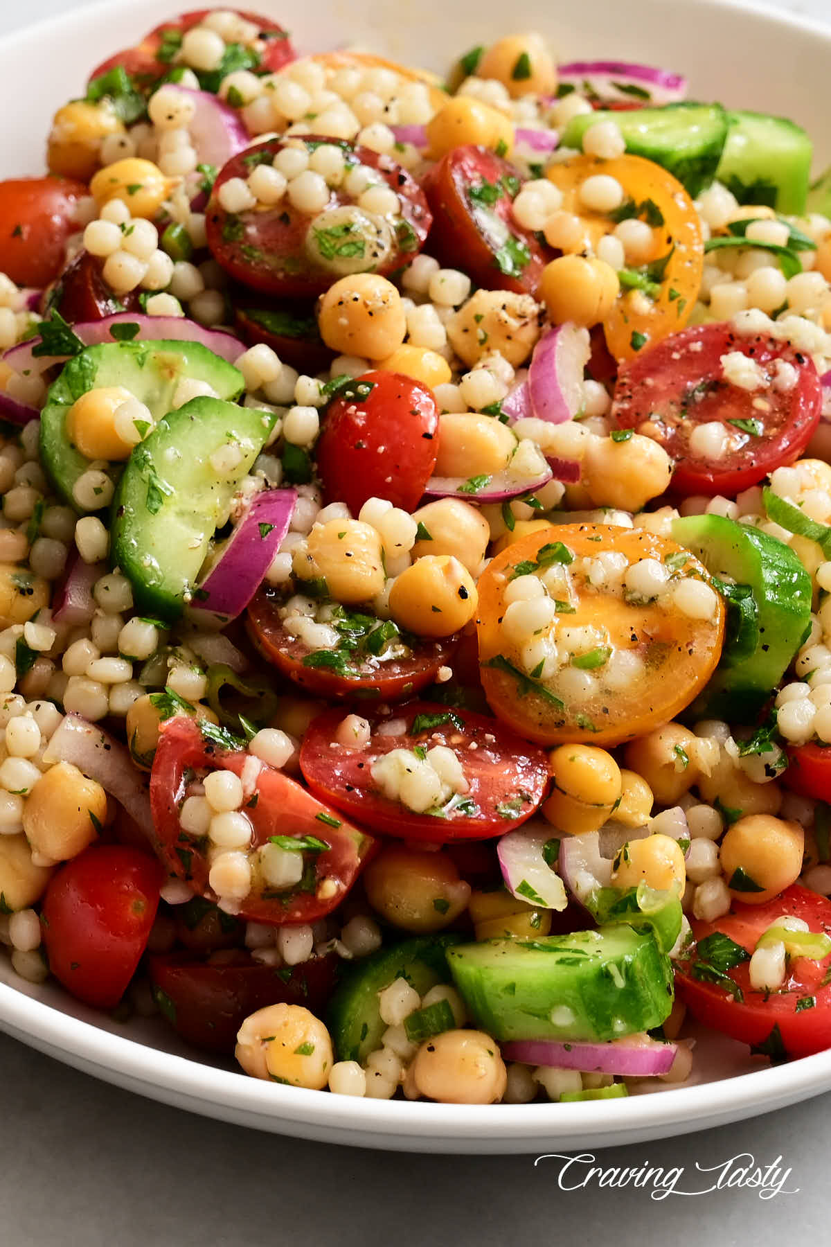 Close up of cherry tomatoes, Israeli couscous, cucumbers, red onion and herbs in a bowl to comprise an Israel couscous salad.