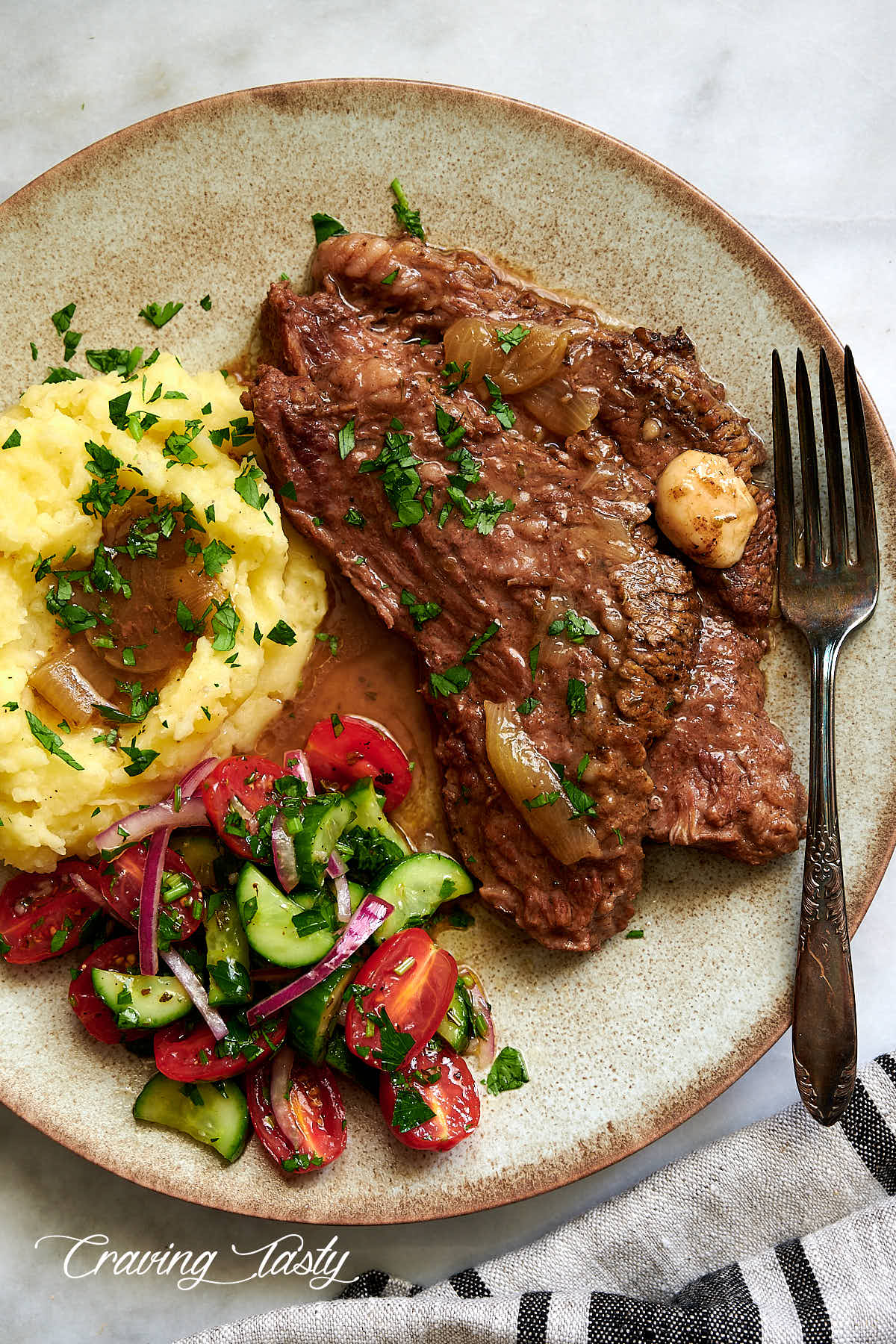 Slices of braised brisket on a speckled dining plate with mashed potatoes and tomato salad.