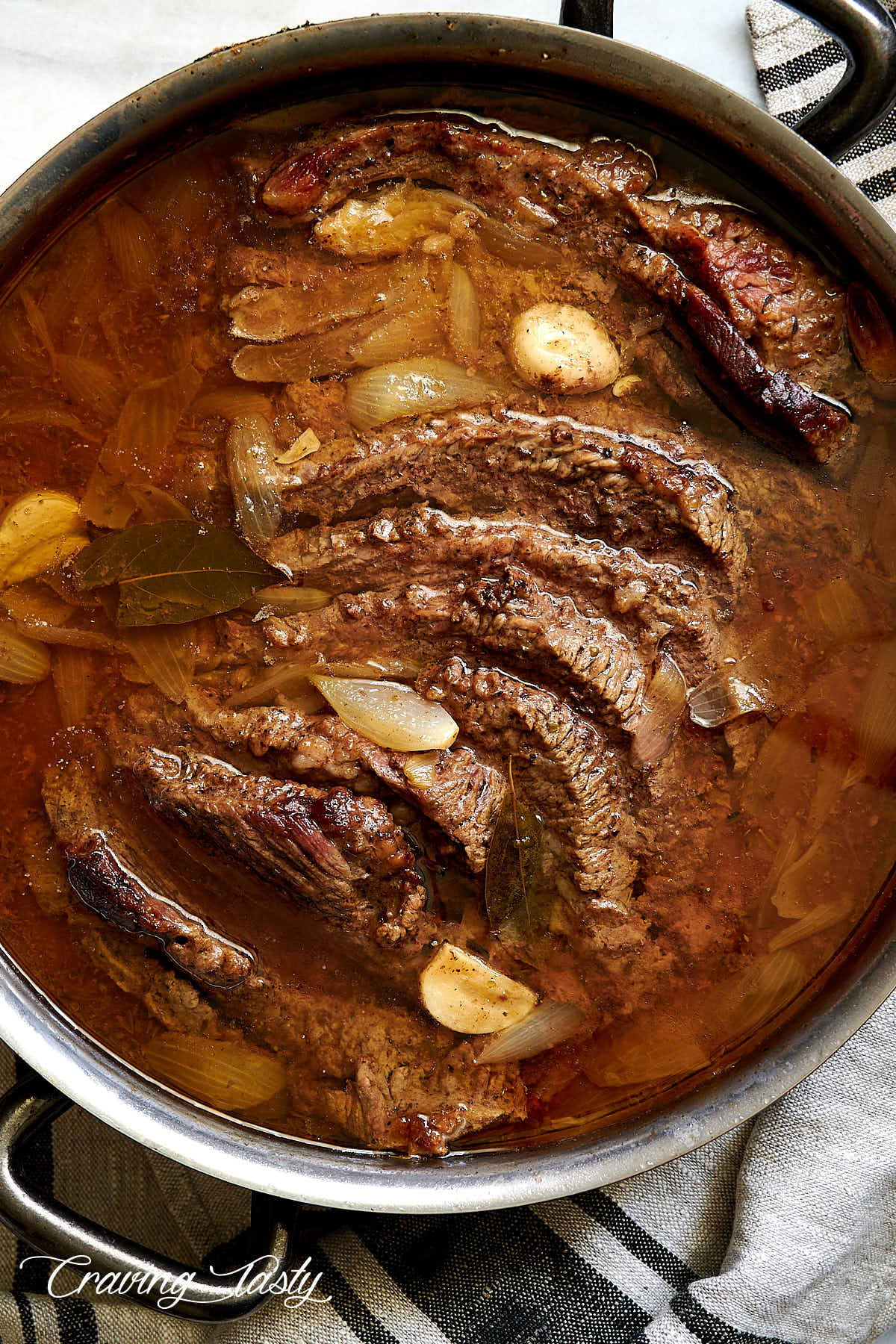 Braised brisket inside a braising pot, sliced, covered with juices, onions and garlic.