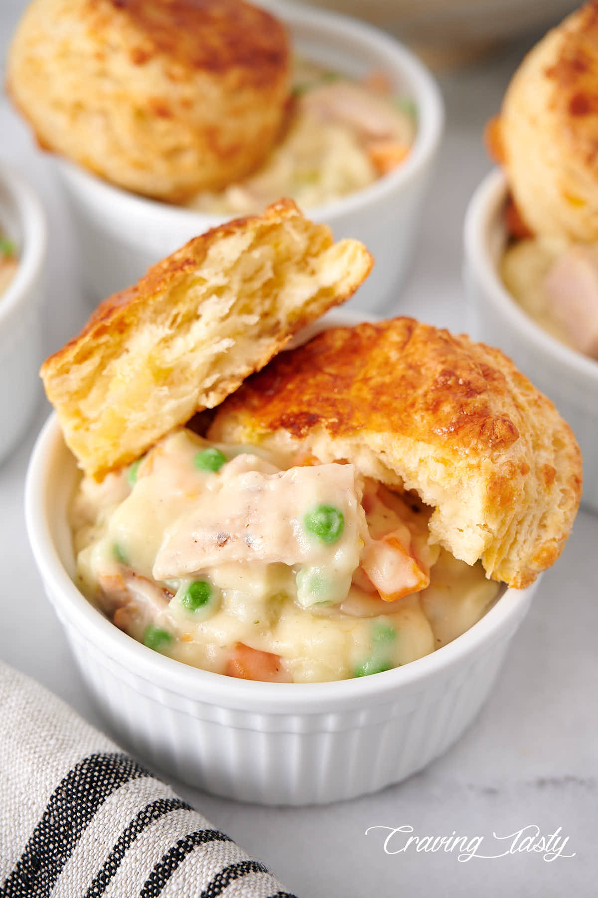 Close up of a bowl of creamy chicken with potatoes, peas and carrots, with biscuit split in half on top.