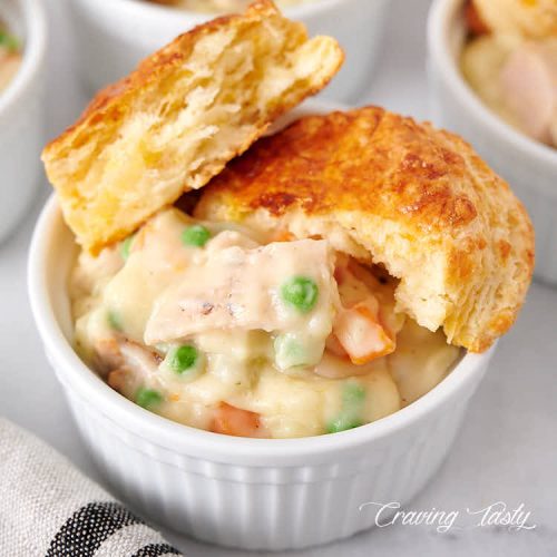 Close up of a bowl of creamy chicken with potatoes, peas and carrots, with biscuit split in half on top.